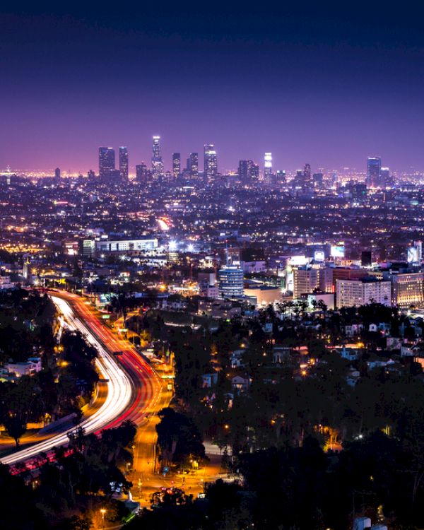 A nighttime cityscape features a brightly lit downtown skyline with tall buildings and a winding highway with busy traffic, surrounded by city lights.