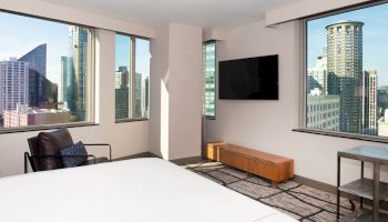 A modern hotel room with a bed, chair, and TV, offering panoramic city views through large windows.