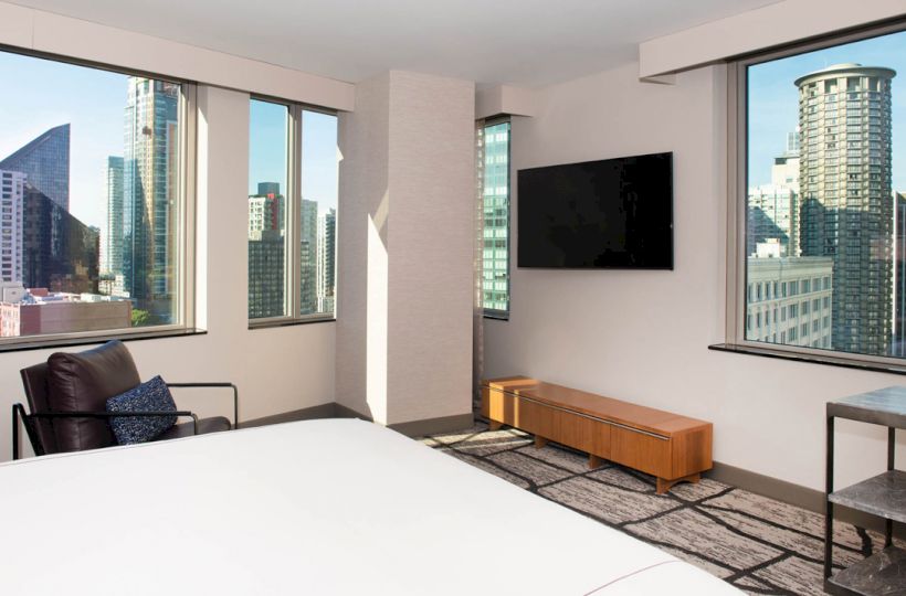 A modern hotel room with a bed, chair, and TV, offering panoramic city views through large windows.