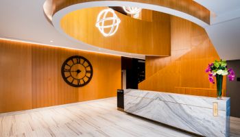 The image shows a modern reception area with a marble counter, floral arrangement, wooden wall paneling, a large wall clock, and decorative lighting.