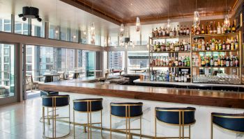 The image shows a modern bar with a long counter, blue and gold bar stools, a well-stocked liquor shelf, and large windows offering city views.