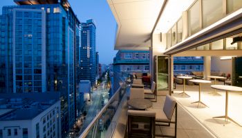 A modern balcony with tables and chairs, overlooking a city street with high-rise buildings at dusk. The area is well-lit and inviting.