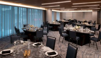 An elegantly set dining room features round tables with black chairs, white napkins, and floral centerpieces, illuminated by modern ceiling lights.