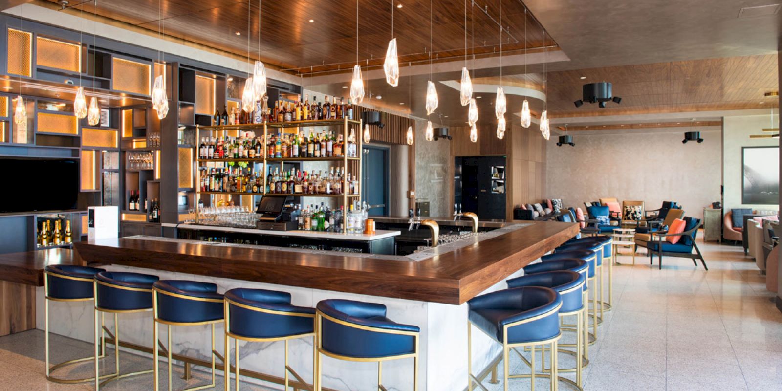 The image shows a modern bar with a wooden countertop, blue and gold bar stools, stylish lighting, and shelves stocked with various bottles of alcohol.