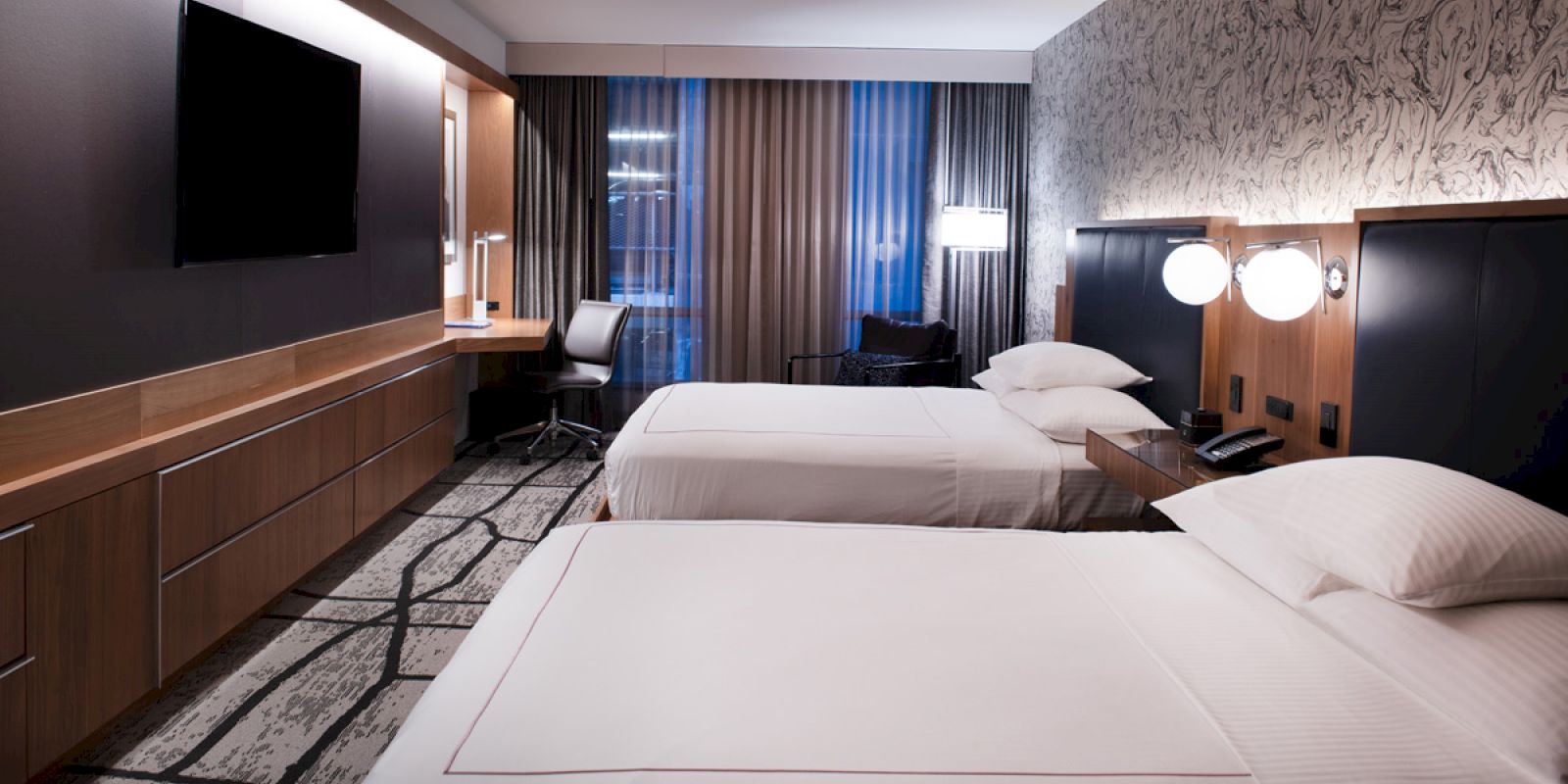 A modern hotel room with two beds, a desk and chair, a wall-mounted TV, and stylish decor.