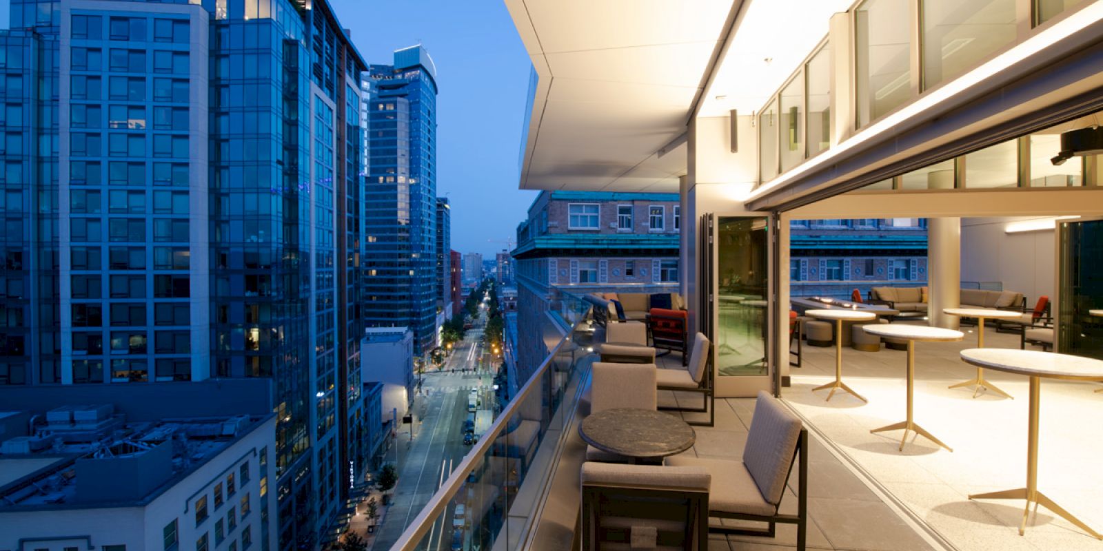 A modern balcony with seating and tables overlooks a street and cityscape with tall buildings at dusk, while interior lights illuminate the space.