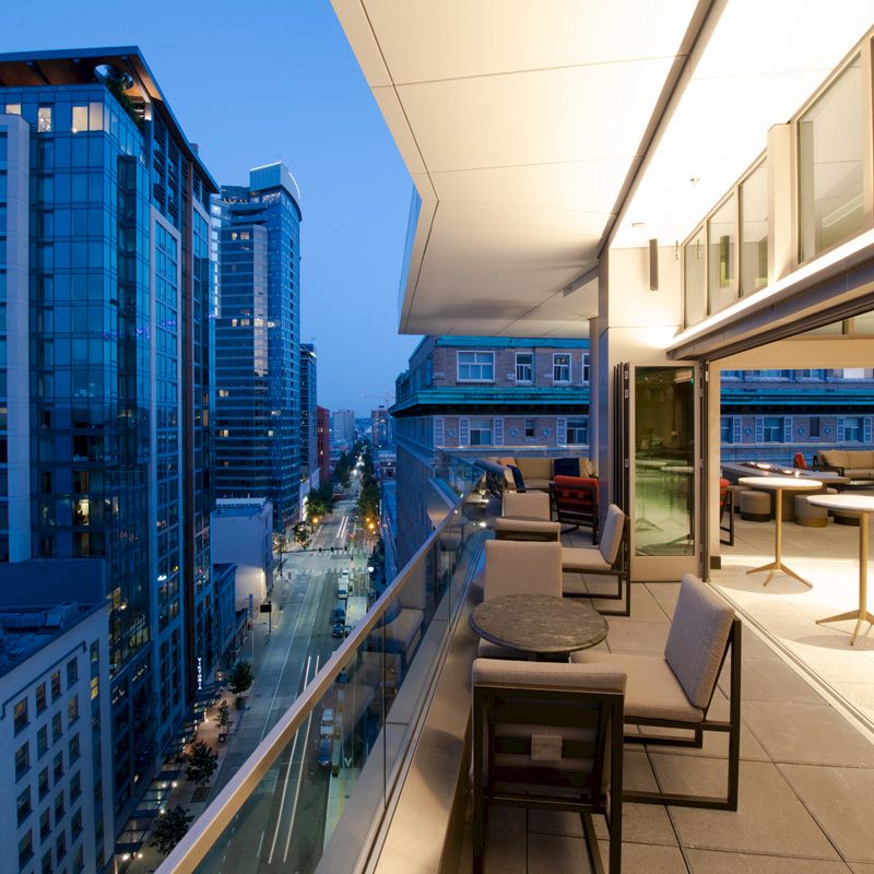 A modern cityscape at dusk with tall buildings and a balcony featuring tables and chairs, overlooking a busy street below.