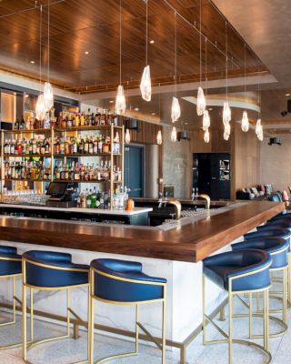 A modern bar with a well-stocked liquor shelf, blue bar stools, pendant lighting, and additional lounge seating is shown in the spacious area.