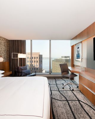 A modern hotel room with a large bed, desk, chair, TV, and a window offering a cityscape view with buildings and the water beyond.