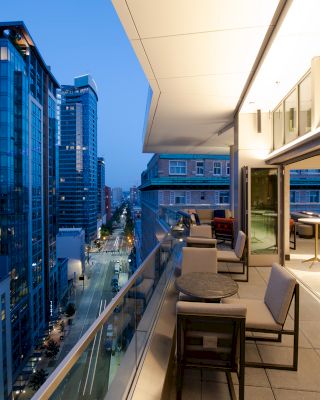 An image of an urban balcony at dusk, overlooking a cityscape with tall buildings and a few outdoor tables and chairs in the terrace area.