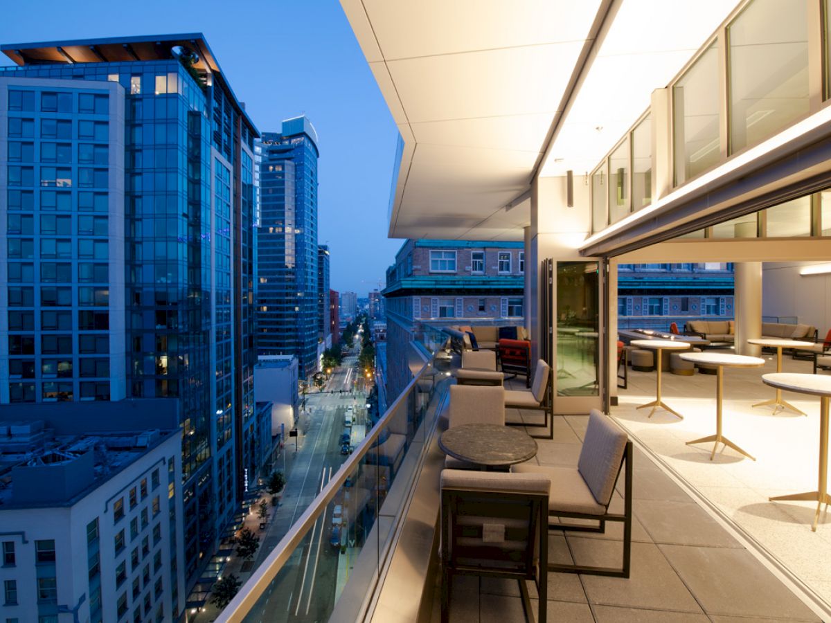 A modern cityscape view from a balcony at dusk, featuring outdoor seating and tables with tall buildings and a street below illuminated by lights.