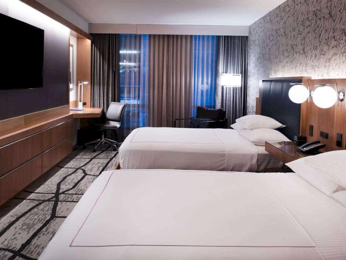 A modern hotel room with two neatly made beds, a flat-screen TV, a desk with a chair, and wall-mounted lamps. The window has curtains.