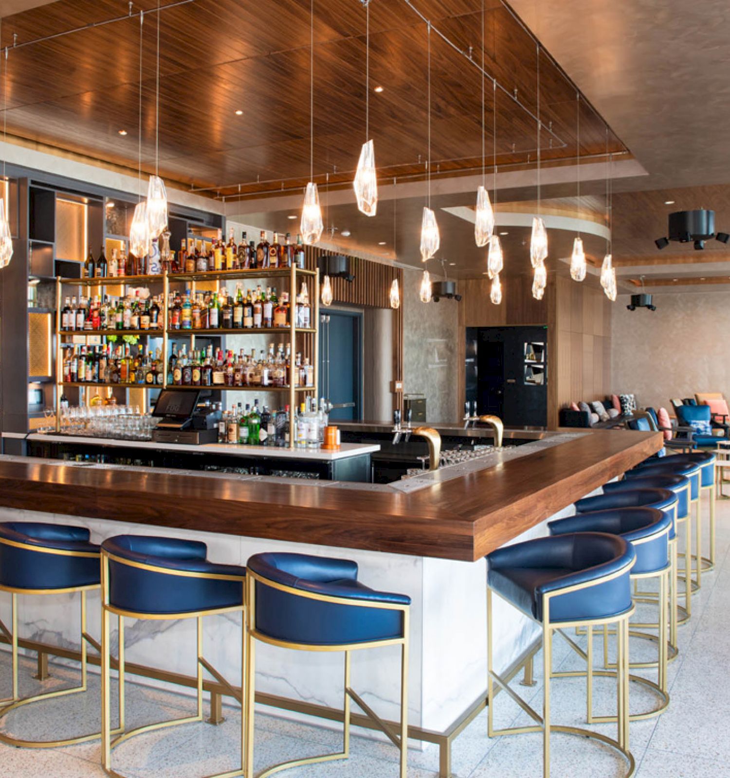 A modern bar with blue stools, a wooden counter, and a well-stocked liquor shelf, surrounded by contemporary decor and lounge seating areas.