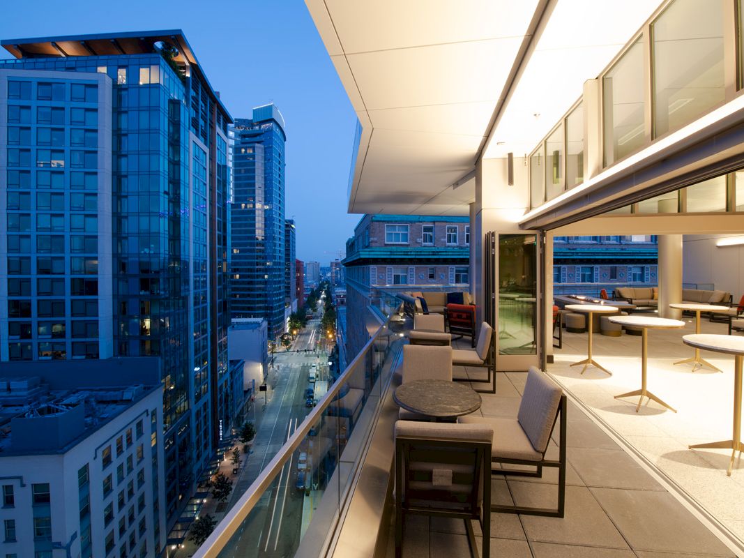 The image shows a modern cityscape viewed from a balcony at dusk. The balcony has outdoor seating and tables, overlooking tall buildings and a street.