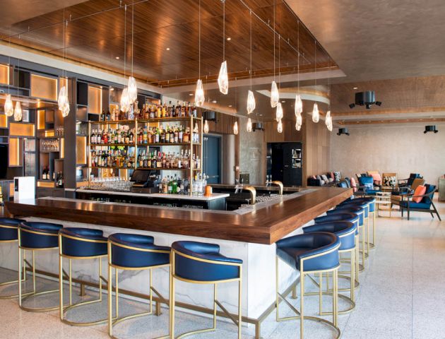 The image shows a modern bar with a wooden counter, blue chairs, numerous hanging lights, and a well-stocked bar shelf in a spacious, stylish setting.
