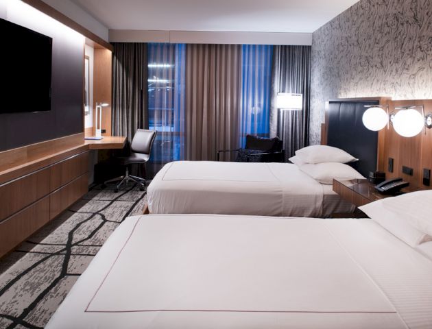 The image shows a modern hotel room with two neatly made beds, a flat-screen TV, a desk, and contemporary decor with a patterned wall.