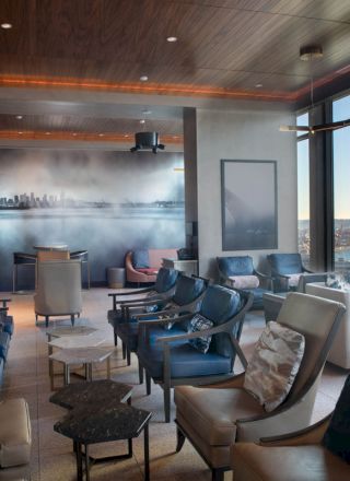 A modern lounge with multiple chairs and tables, large windows offering a view of the water and a Ferris wheel in the distance, is shown here.