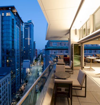 A modern urban balcony during twilight with tables and chairs, offering a cityscape view of buildings and a street below.