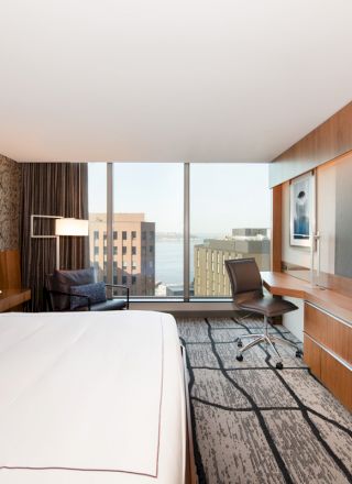 A modern hotel room with a large bed, a wall-mounted TV, a desk and chair, a window with a city view, and stylish decor.