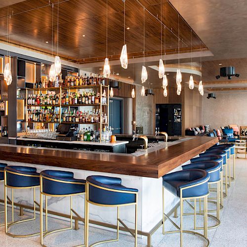 The image shows a modern bar with a large counter, blue barstools, hanging lights, and a well-stocked liquor shelf. There's seating in the back.