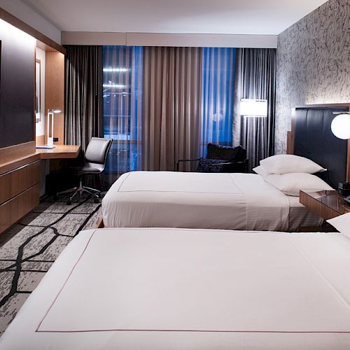 The image shows a modern hotel room with two neatly made beds, a flat-screen TV, a desk with a chair, and contemporary decor.