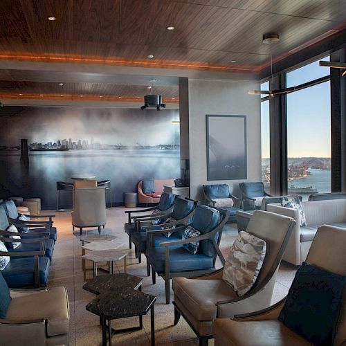A modern lounge area with leather chairs, large windows overlooking a cityscape and a body of water, and contemporary wall art.