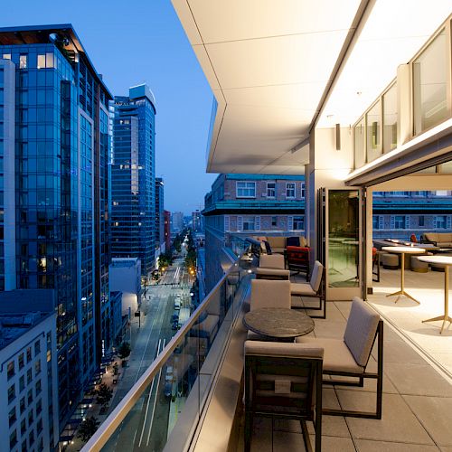 A modern balcony overlooking a cityscape at dusk, with outdoor seating and tables, adjacent buildings, and a view down a city street.
