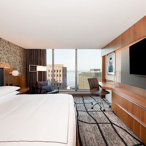 A modern hotel room with a large bed, flat-screen TV, desk, and a view of buildings and water through floor-to-ceiling windows.