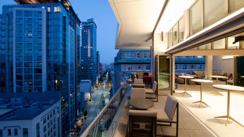 A modern balcony at dusk with tables and chairs, overlooking a city skyline with tall buildings and a busy street below.