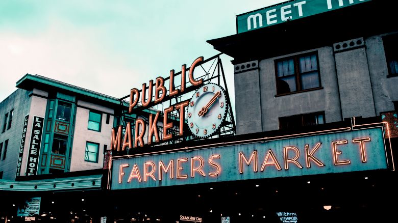 Image depicts a neon sign at a urban Public Market, marking a Farmers Market. There are nearby buildings with visible signs and a clock.