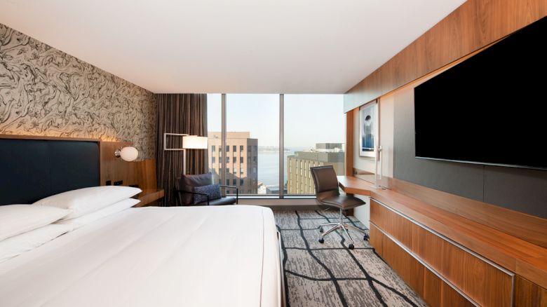 A modern hotel room features a large bed, wall-mounted TV, desk with chair, and a view of buildings and water through large windows.