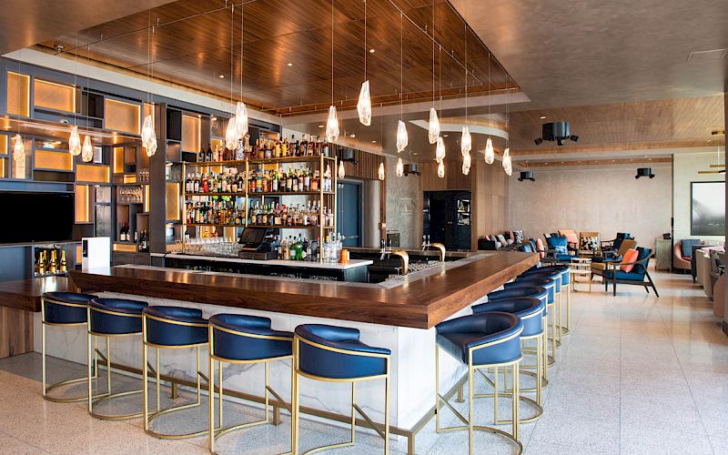 A modern bar with a central counter, high chairs, hanging lights, and a well-stocked shelf of liquors and spirits.
