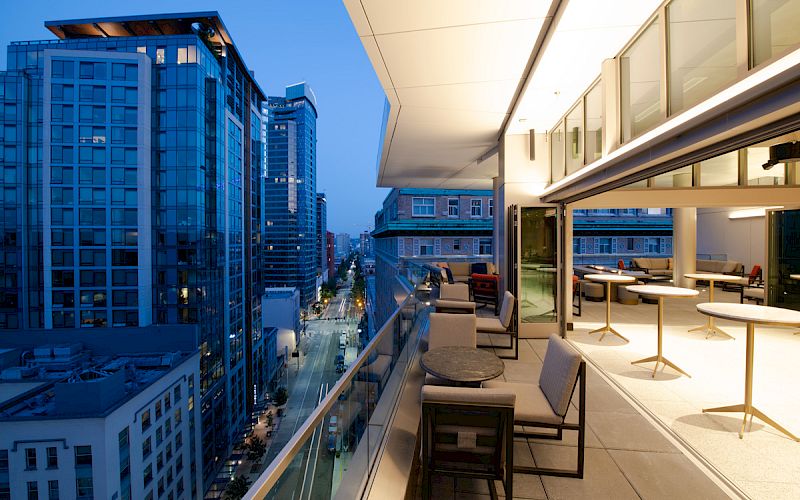 The image shows a modern urban balcony with outdoor seating, overlooking a cityscape with tall buildings at dusk.