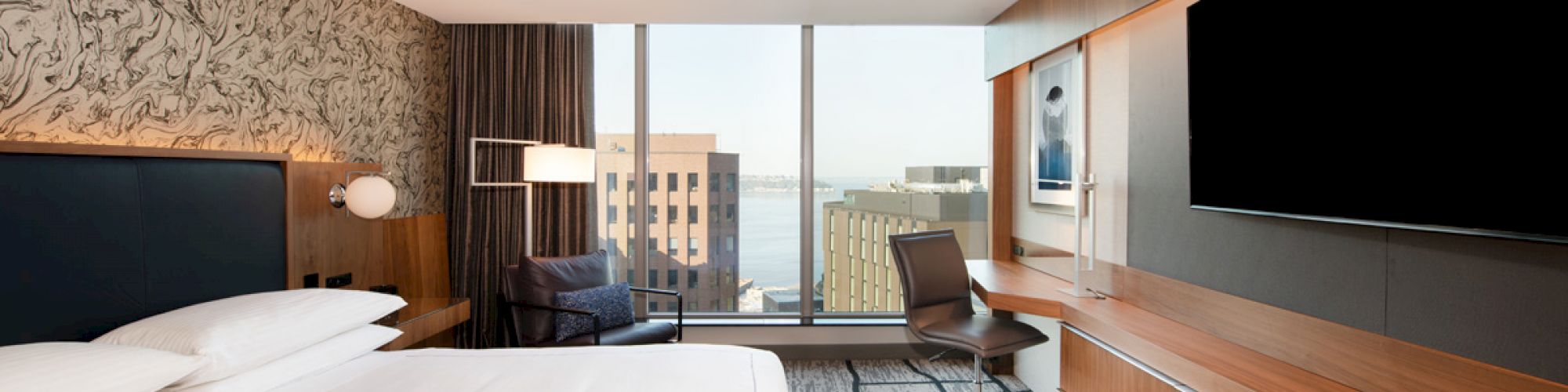 A modern hotel room with a large bed, a desk, a chair, a TV, and a large window offering a view of nearby buildings and the water.