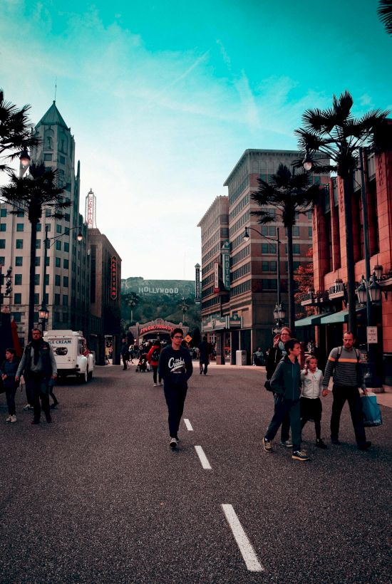 People walking down a street with tall buildings on either side, palm trees, and a sign in the background resembling the Hollywood sign.