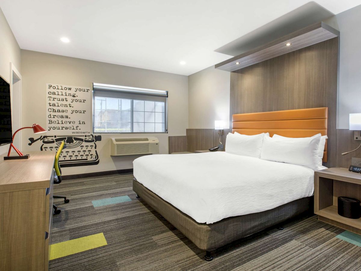 A modern hotel room with a king-size bed, desk, office chair, mounted TV, wall art of a typewriter, and a motivational quote on the wall.