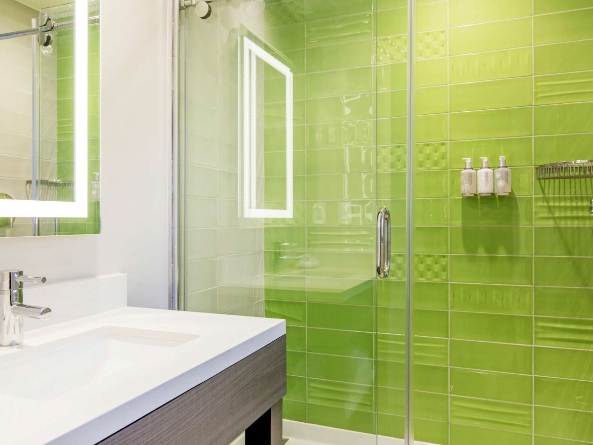 The image shows a modern bathroom with a bright green tiled shower, glass door, and a white sink with a mirror.